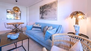 A living room with a blue sofa, a round wooden coffee table, a wicker chair, a wall-mounted painting, and a floor lamp. - Sunny 2-Bedroom Apartment | Pool & Terrace | Estepona (Estepona)