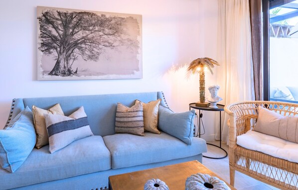 Een woonkamer met een bank, een bijzettafeltje, een lamp en een rieten stoel.