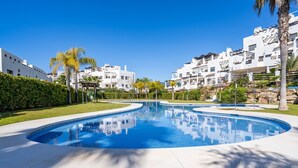 A swimming pool with a curved edge, surrounded by a manicured lawn and palm trees, with white buildings in the background. - Sunny 2-Bedroom Apartment | Pool & Terrace | Estepona (Estepona)