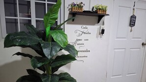 A room with a large plant, a white door, a wall with a welcome message, and a small shelf with flowers. - Apartamento Lilia Airport 1 (Panamá)