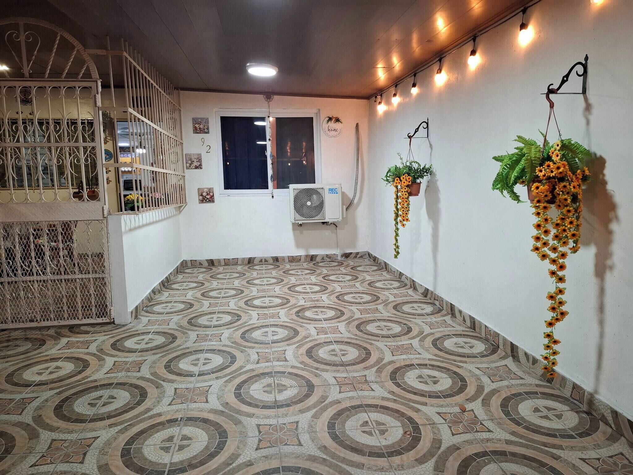 A room with patterned floor tiles, a window with curtains, a wall-mounted air conditioner, and hanging plants.