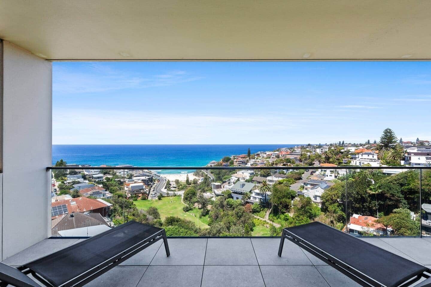 A balcony with two black lounge chairs, a clear view of the ocean, and a residential area in the background.