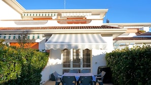 Eine Terrasse mit Tisch und Stühlen, ein überdachter Bereich mit gestreifter Markise und ein weißes Gebäude mit einem Ziegeldach.