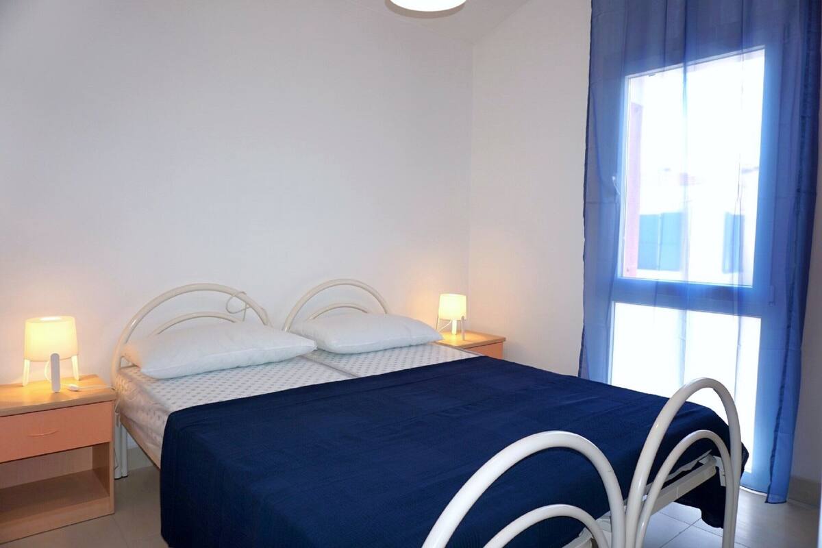 A bedroom with a bed, two bedside tables with lamps, a window with curtains, and a view of the outside.