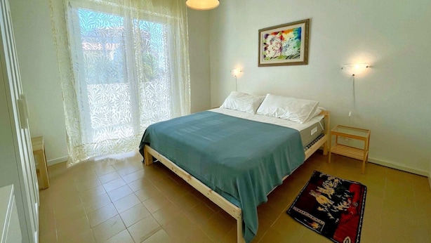 Un dormitorio con cama, una ventana con cortinas translúcidas, un cuadro colgado en la pared y una mesita de noche con lámpara.
