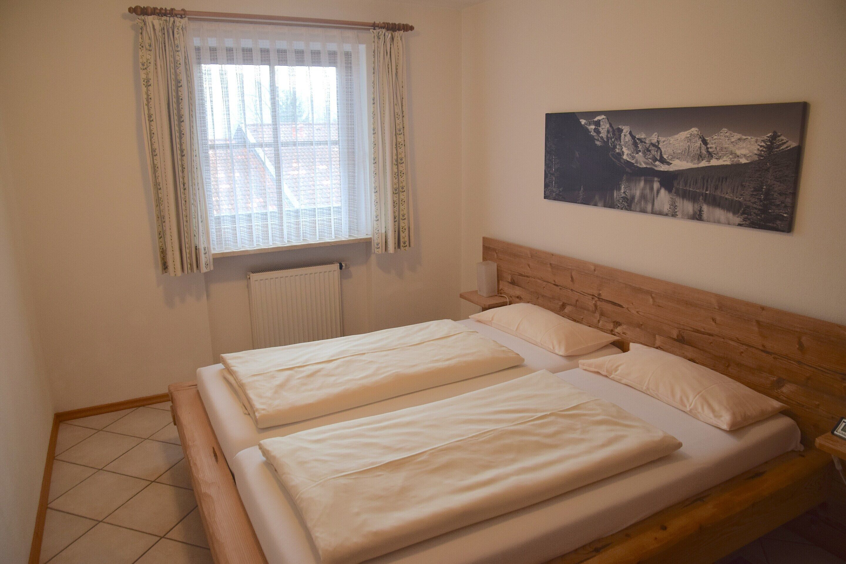 Ein Schlafzimmer mit zwei Betten, einem fensterbelaubten Fenster, einem Heizkörper und einem Wandbild von einer Berglandschaft.