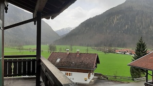 A mountain view from a balcony with a wooden railing, a white building with a red roof, and a green field. - Beautiful Lodging in the Alps Near Bayrischzell (Bayrischzell)
