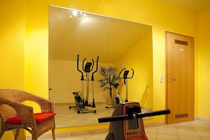 Una habitación con un espejo grande que refleja equipo de ejercicio, una silla de mimbre con un cojín rojo y una puerta de madera.