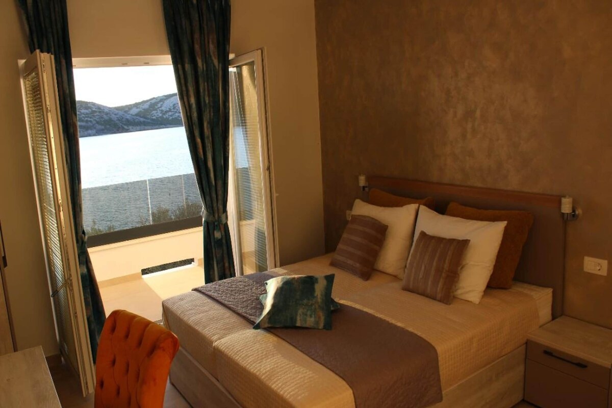 Een slaapkamer met een bed, kussens, een nachtkastje, een stoel en uitzicht op zee door een raam.