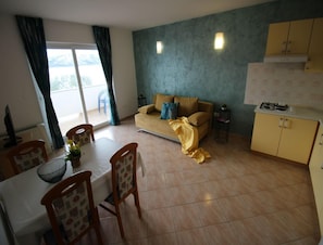 A living room with a sofa, a dining table with chairs, a kitchen area with cabinets, and a view through a window. - Comfortable Croatian Couple Retreat (Novalja)