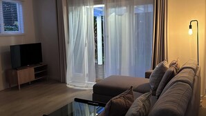 A living room with a sofa, a TV, a glass coffee table, sheer curtains, and a lamp. - Central & Cozy Raglan house - 10min walk to town! (Raglan)