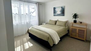 A bedroom with a bed, a bedside table with a plant, a painting on the wall, and a window with curtains. - Central & Cozy Raglan house - 10min walk to town! (Raglan)