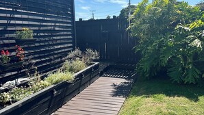 A wooden deck with a grassy area, a metal fence, and a building with a sloped roof. - Central & Cozy Raglan house - 10min walk to town! (Raglan)