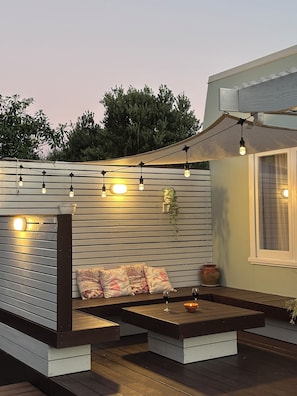 A wooden deck with a built-in seating area, string lights, and a potted plant. - Central & Cozy Raglan house - 10min walk to town! (Raglan)