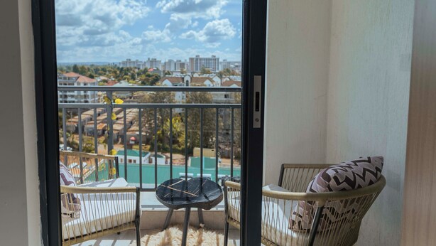 A balcony with wicker furniture, a fur rug, and a view of buildings and greenery. - Studio Apartment with a pool in Ruaka (Ruaka)