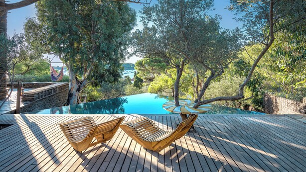 Una piscina infinitat amb un pont de fusta, envoltada d'arbres i una vista al mar.