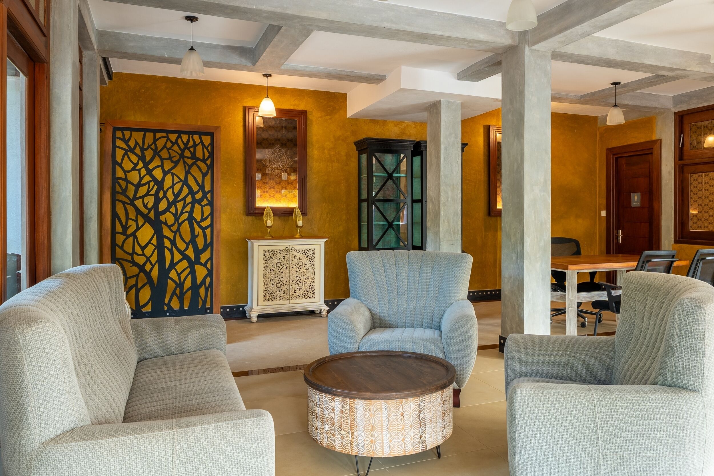 A modern lobby with a round wooden coffee table, two grey armchairs, a decorative wall panel, and a wooden cabinet.