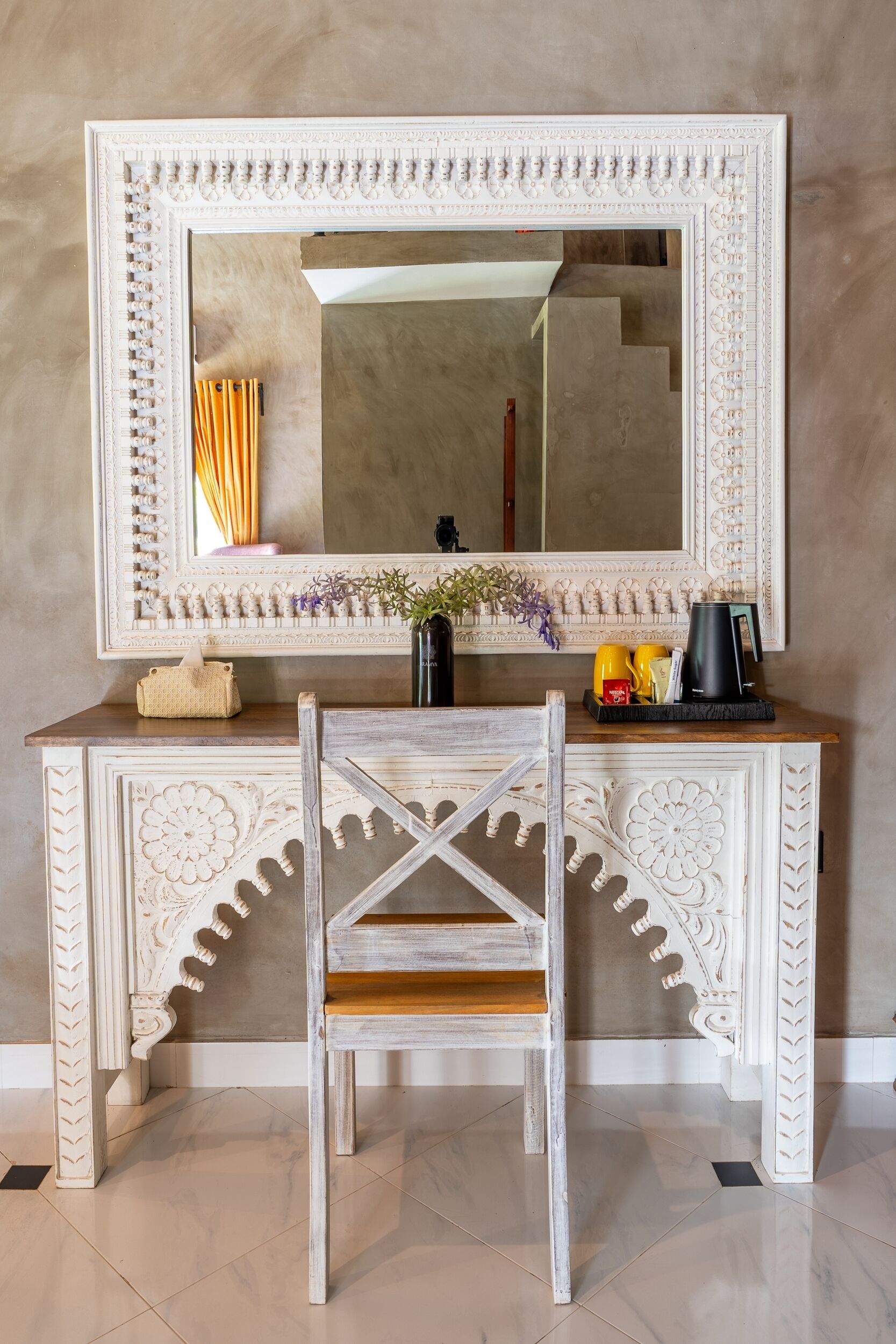 A white ornate console table with a mirror above it, a chair in front, and a tray with cups and a coffee maker.