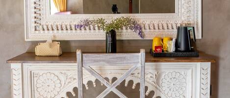 A white ornate console table with a mirror above it, a chair in front, and a tray with cups and a coffee maker.