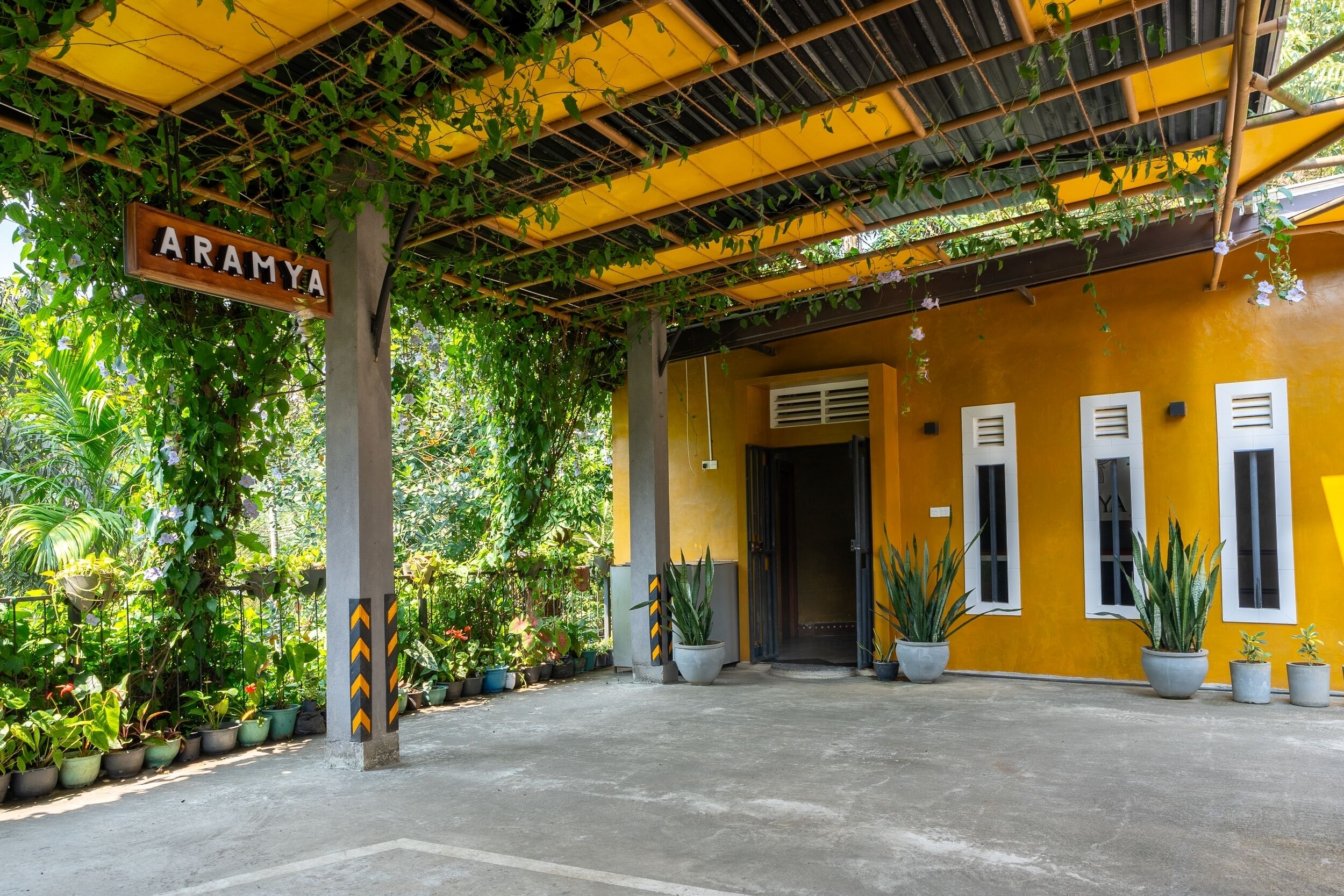 A covered entrance with a sign reading 'ARAMYA,' potted plants, and a concrete floor.