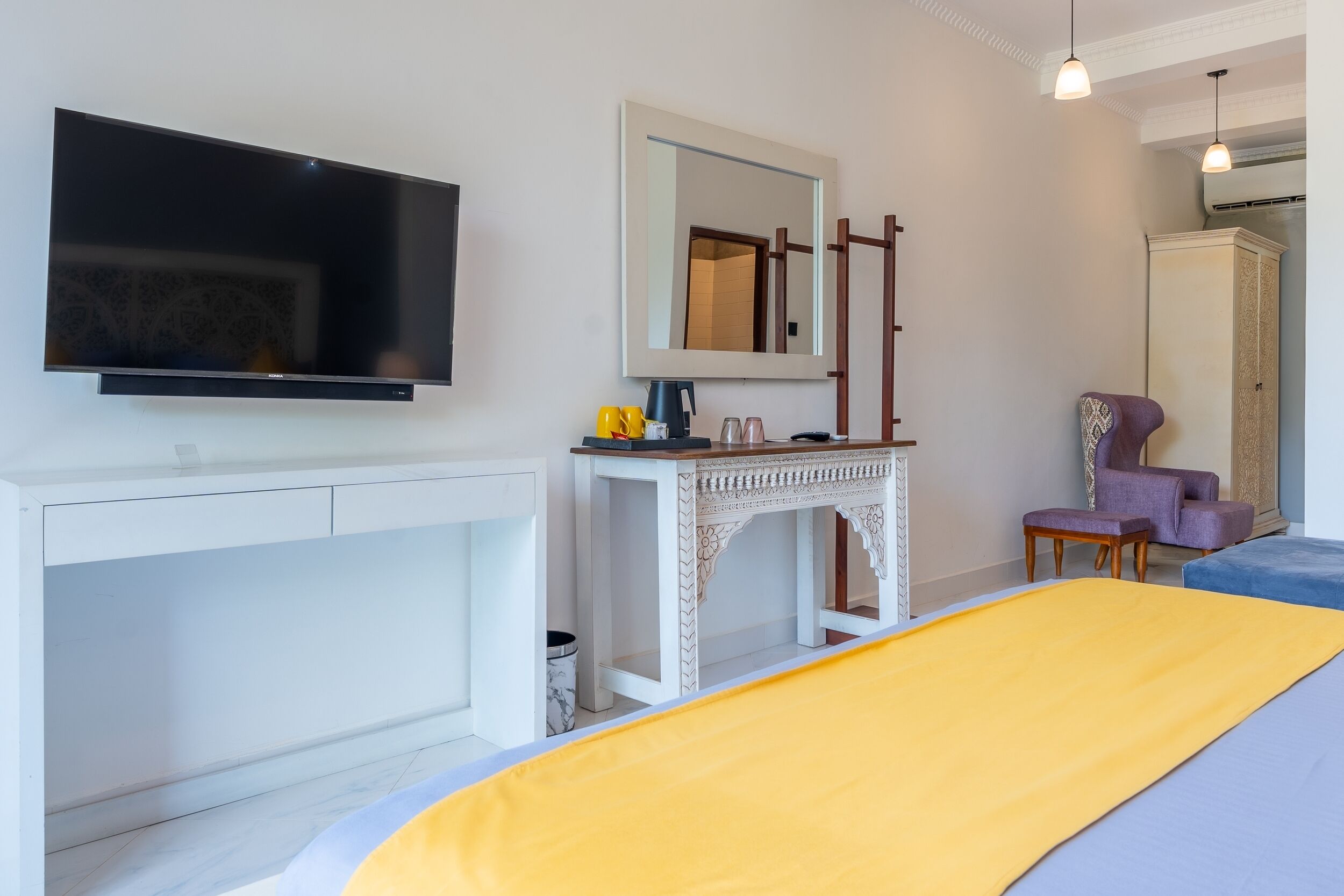Une chambre d’hôtel moderne dotée d’une télévision à écran plat, d’une console blanche, d’un grand lit recouvert d’une couvre-lit jaune et bleue, d’un fauteuil violet et d’un grand miroir.
