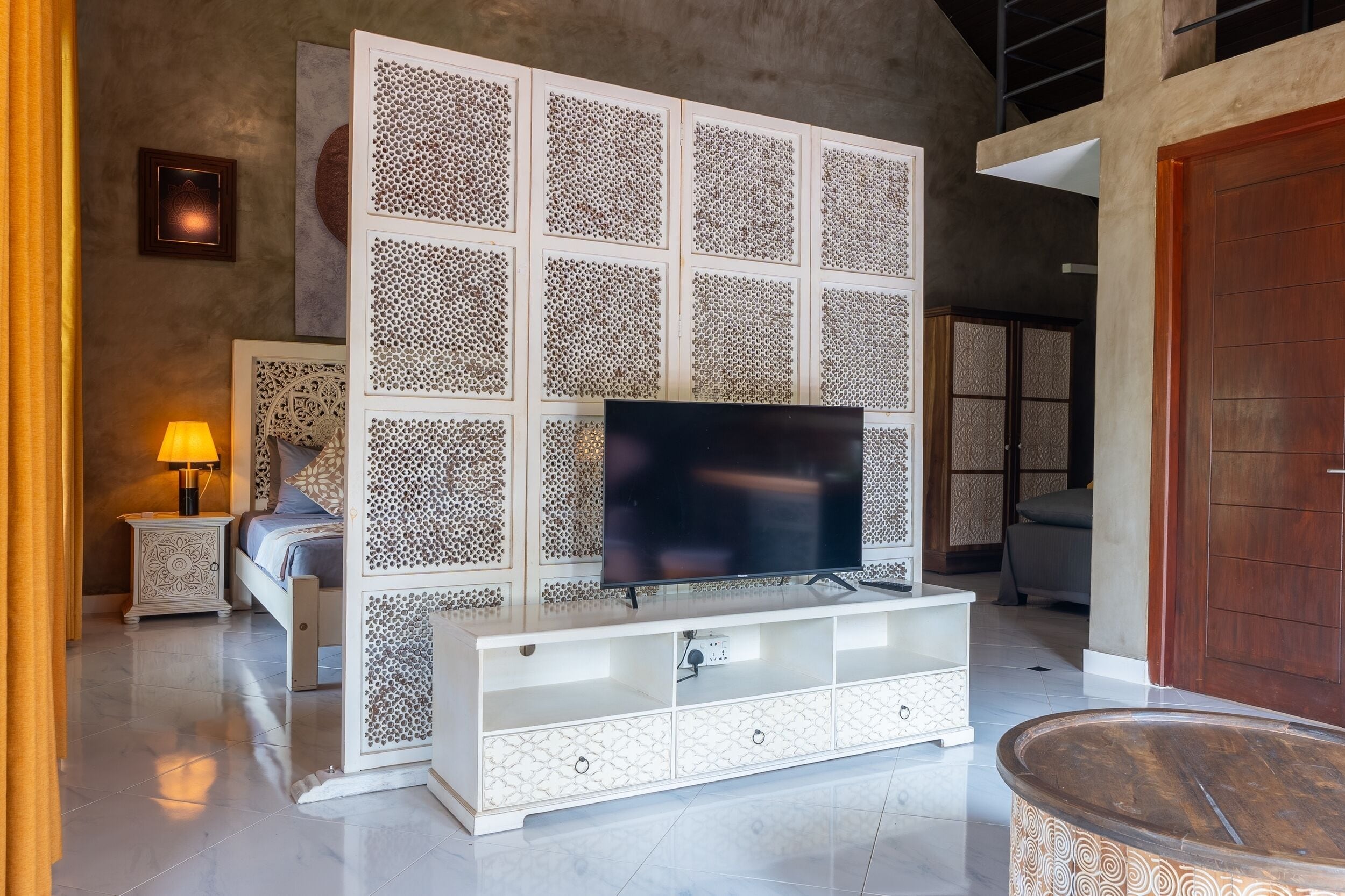 Uma sala de estar moderna com uma televisão em um armário branco, uma cama com cabeceira estampada e uma porta de madeira.