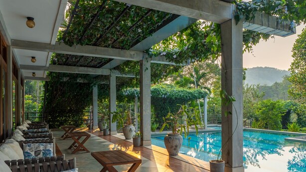 A covered outdoor seating area with a pool, wooden furniture, and potted plants. - Aramya Riverfront Boutique Resort (Kitulgala)