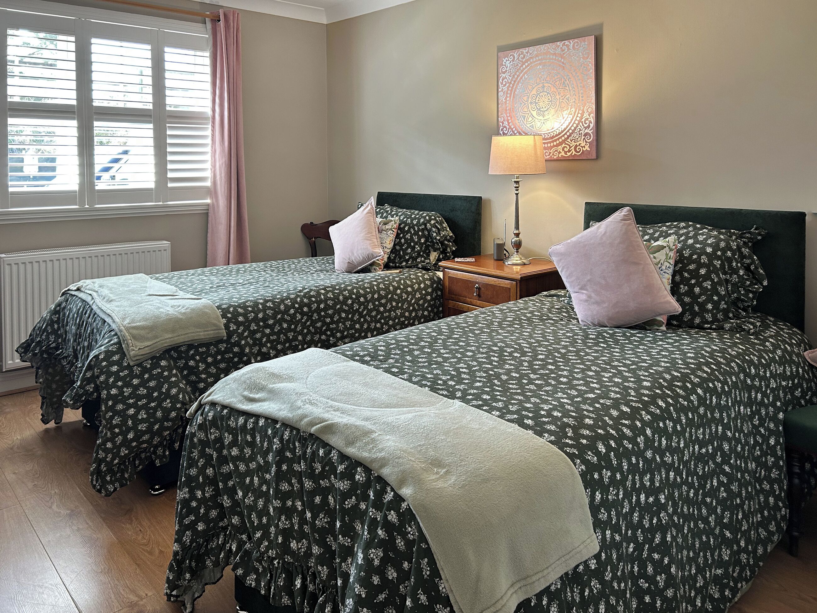 A room with two beds, a nightstand, a lamp, a radiator, and a window with blinds.
