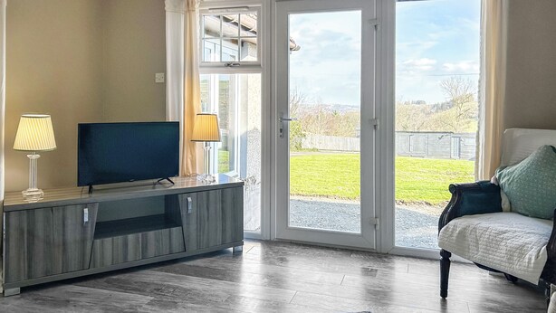 Uma sala de estar com televisão, abajur, sofá e vista para uma paisagem verde através de uma porta de vidro.