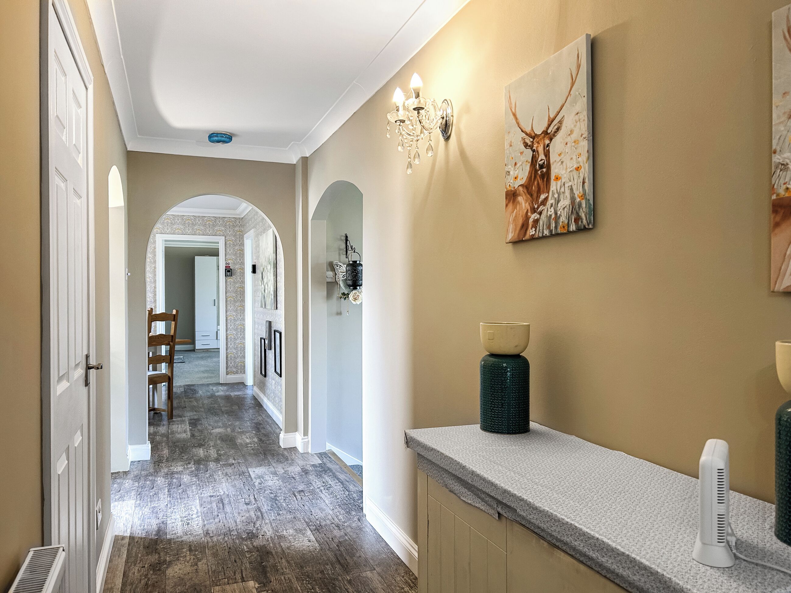 A hallway with a wooden floor, a white door, a chandelier, and a deer painting.