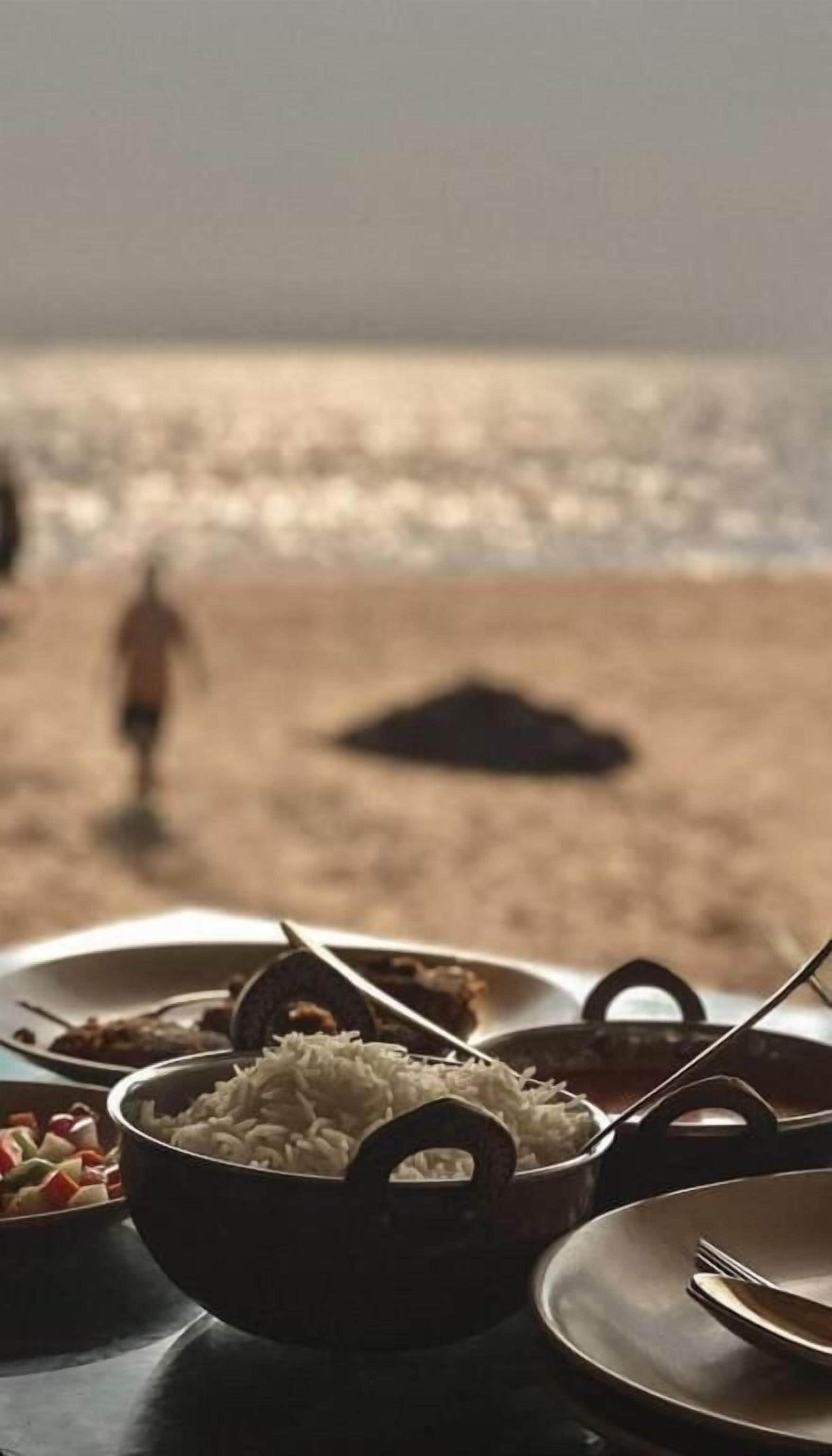 Una mesa con un bol de arroz, un plato de comida y una cuchara. Al fondo, se ve a una persona caminando en la playa y una duna de arena.