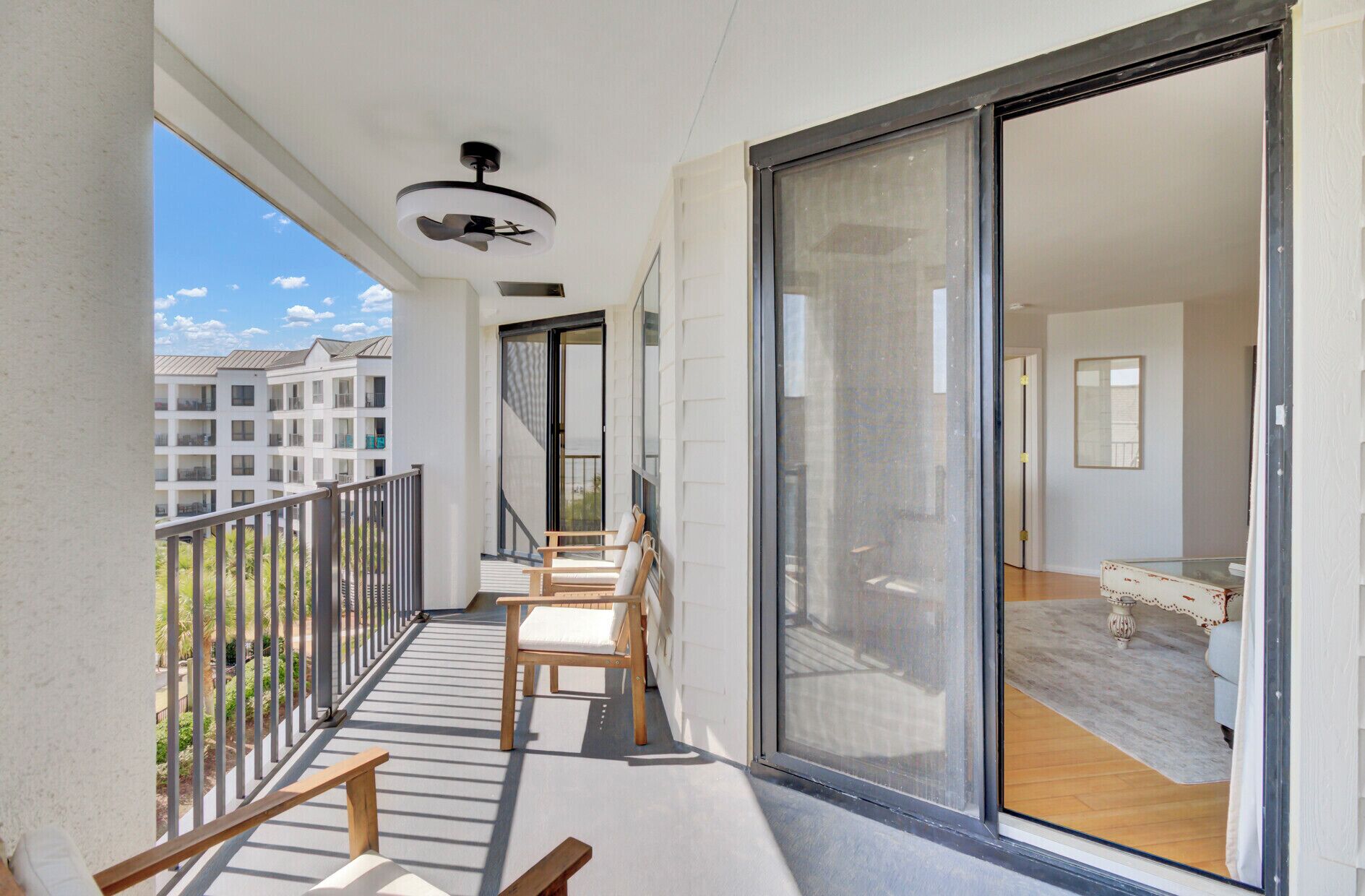 A balcony with a ceiling fan, a sliding glass door leading to a room with a bed and a small table, and a view of other buildings.