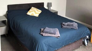 Een netjes opgemaakt bed met een donkerblauwe sprei en twee gevouwen handdoeken, een bedlampje en een klein nachtkastje.