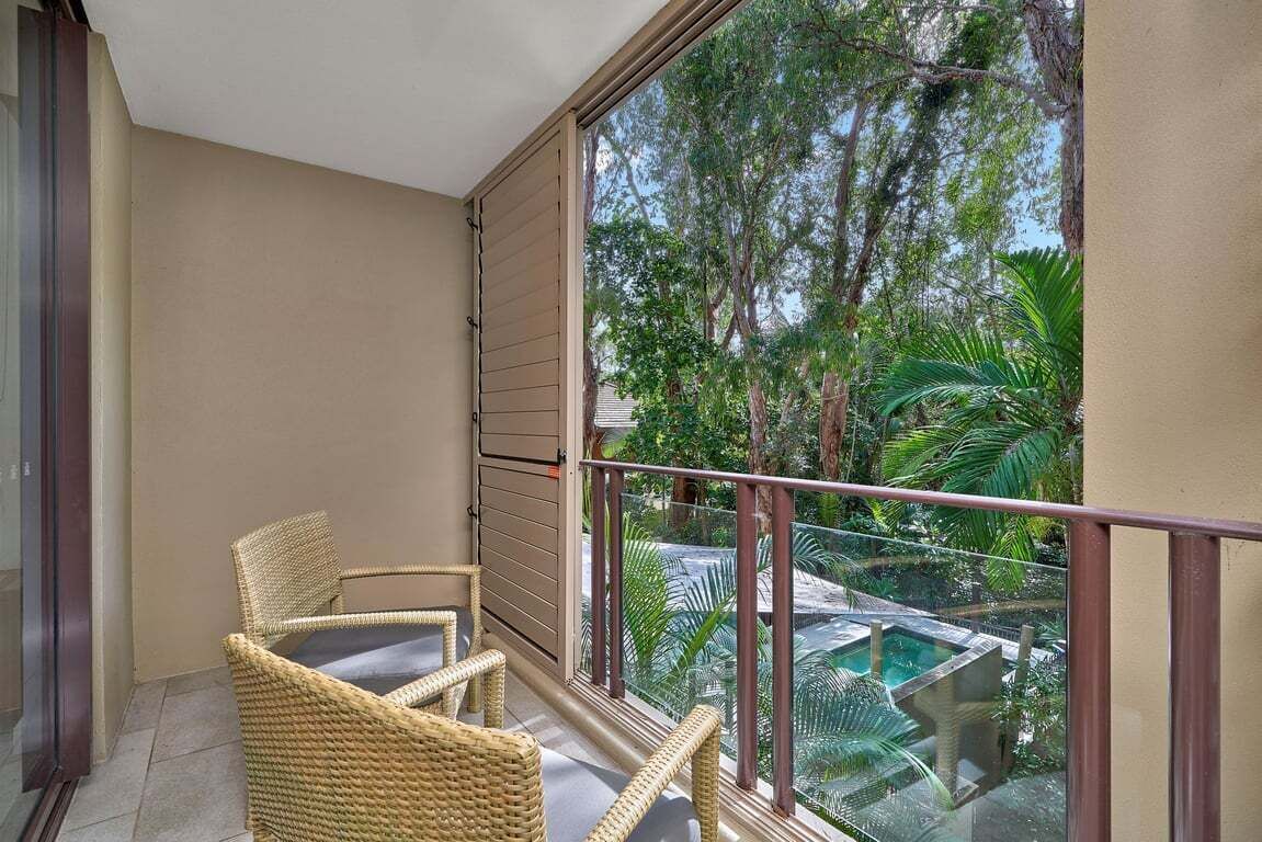 Un balcon avec des fauteuils en osier, une balustrade en verre et une vue sur une piscine et des arbres.