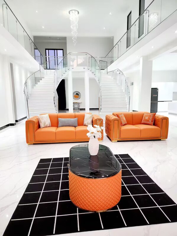 A modern living room with two orange sofas, a glass coffee table, and a staircase with glass railings.