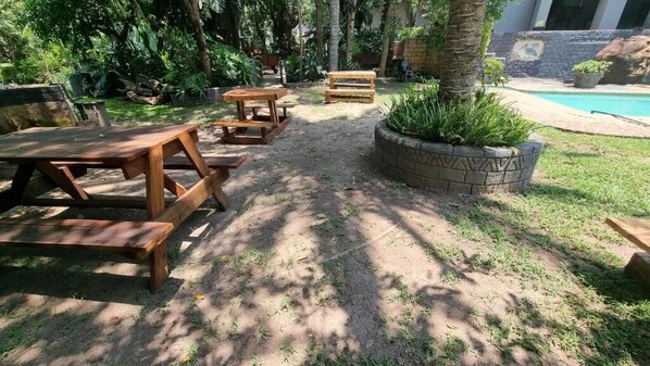An outdoor patio with wooden tables and benches, surrounded by greenery and a pool.