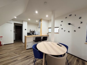 Ein modernes Hotelzimmer mit Holzboden, einem runden Tisch mit Stühlen, einer Küchenzeile und einer Wanduhr.