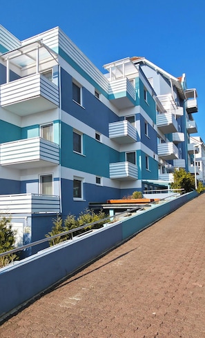 Un immeuble résidentiel à plusieurs étages, doté de balcons, d’un chemin pavé et surplombant un ciel bleu dégagé.