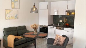 Ett modernt vardagsrum med en grön soffa, ett litet runt bord med en vas, en grå fåtölj och en köksdel med vita skåp och en svart bänkskiva.