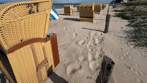 Flätade strandstolar på en sandstrand, med fotspår, en flagga och en trästolpe.