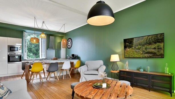 A modern kitchen with a dining area, a round wooden table, and a green wall with a painting.