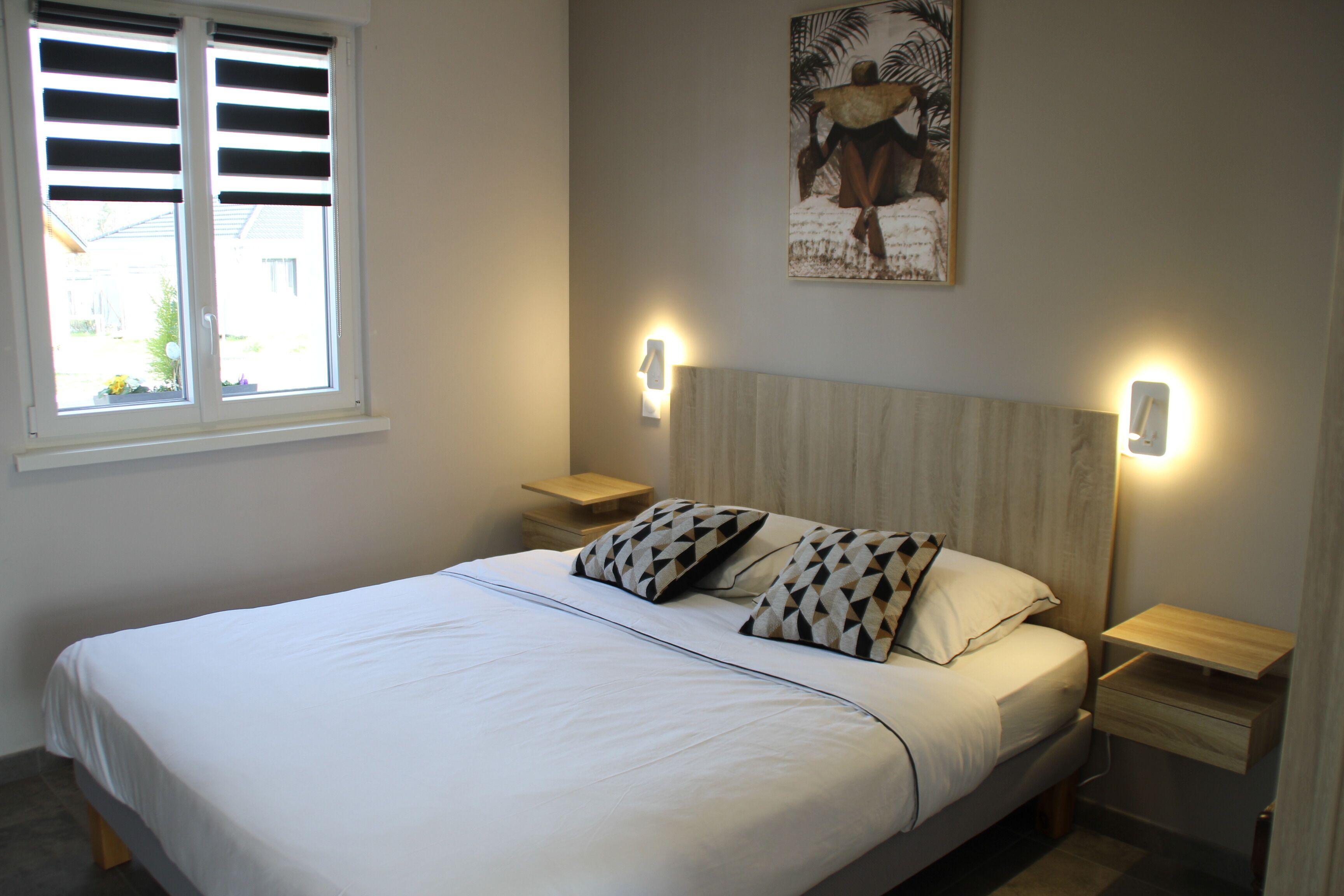 Une chambre à coucher comprenant un lit, deux tables de chevet, une fenêtre avec des stores, un tableau accroché au mur et deux lampes murales.