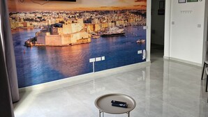 A room with a large cityscape mural, a flat-screen TV mounted on the wall, a small round table, and a tiled floor. - Penthouse Penthouse Green House A27 - Green style (Saint Paul's Bay)