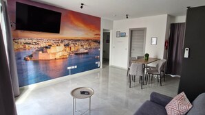 A modern living room with a large TV, a sofa, a small table, and a cityscape mural on the wall. - Penthouse Penthouse Green House A27 (Saint Paul's Bay)
