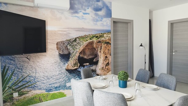 A dining area with a table set for four, a potted plant, and a wall mural of a coastal scene. - Penthouse Penthouse Green House A27 (Saint Paul's Bay)