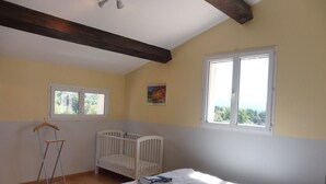 A room with a crib, a bed, a wooden floor, a window, and a ceiling with exposed beams. - Magnificent villa at the foot of Mont Ventoux (Bédoin)