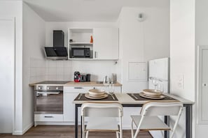A modern kitchen with white cabinetry, a microwave, a stove, and a dining area with two chairs and a table. - Chic and brand-new studio near Paris (Bondy)