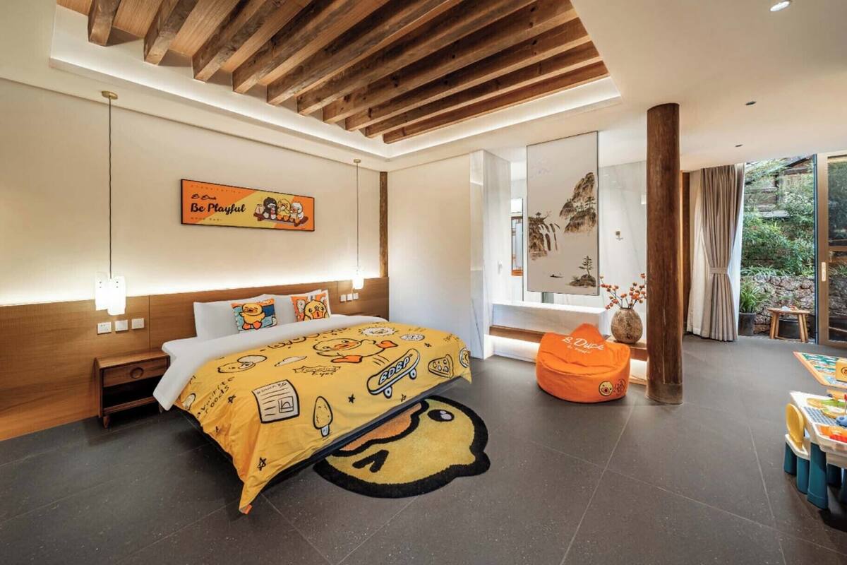 A modern bedroom with a wooden ceiling, a bed with a yellow blanket, a wooden nightstand, a large window, and a view of greenery outside.