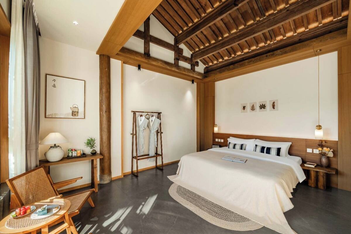 A modern hotel room with a large bed, wooden ceiling beams, a seating area with a table and chairs, and a wall-mounted robe rack.