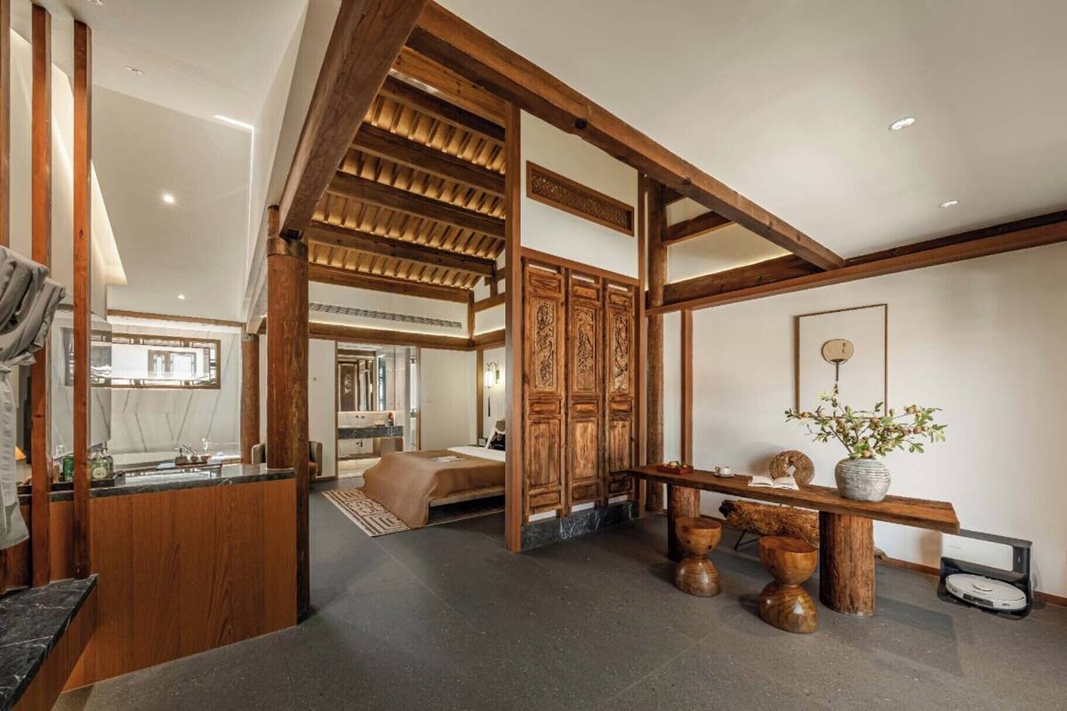 A spacious hotel room with a large bed, wooden beams, a dining area with a table and chairs, and a view of the adjacent bathroom.